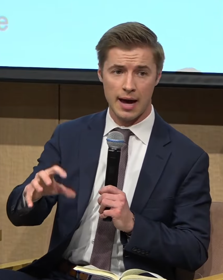 Ryan Fedasiuk speaking into a handheld microphone on a panel.